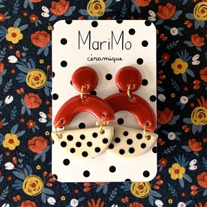 Peut inclure: Boucles d'oreilles en céramique rouges et crème. Elles présentent un cercle rouge en haut, une arche rouge et un demi-cercle crème à pois noirs. Elles sont présentées sur une carte blanche avec le texte "MariMo céramique".
