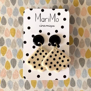 May include: A pair of ceramic earrings with a black and white polka dot design. The earrings have a black circular top and a cream-colored, scalloped-edge bottom with black dots. The earrings are on a white card with the text "MariMo céramique".