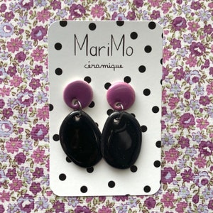 Puede incluir: Un par de pendientes de cerámica con forma ovalada negra y parte superior redonda de color morado. Los pendientes se muestran en una tarjeta blanca con lunares negros y el texto "MariMo céramique".