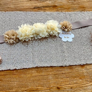 May include: A light brown satin sash with a cluster of ivory and beige fabric flowers. The sash has a white cloud-shaped tag with the text "Mommy to be" in gold lettering.