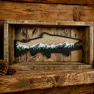 May include: Wooden framed art piece featuring a fish silhouette with a mountain and forest scene. The fish is black with a light brown interior, and the mountains are white. The artwork is set against a rustic wooden background.