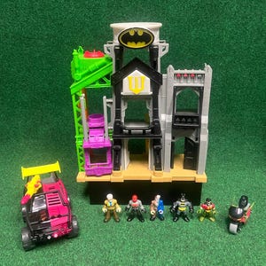 Peut inclure: Un ensemble de jeu sur le thème de Batman comprenant un bâtiment à plusieurs niveaux avec un Bat-Signal, un véhicule rose et noir, une grue verte et plusieurs figurines. L'ensemble est sur une surface verte.
