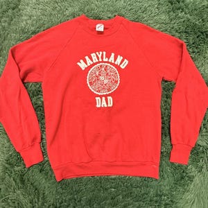 Op de afbeelding: Een rode sweatshirt met lange mouwen. De voorkant heeft een wit cirkelvormig ontwerp met het woord "MARYLAND" bovenaan, een universiteitszegel in het midden en het woord "DAD" eronder. De sweatshirt is gemaakt van zacht materiaal.