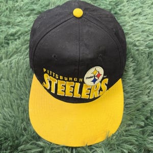 Puede incluir: Una gorra de béisbol negra con visera amarilla y un botón amarillo en la parte superior. El frente presenta el texto bordado Pittsburgh Steelers en letras amarillas, acompañado del logotipo circular de los Steelers con formas de diamante amarillas, rojas y azules.