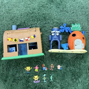 Puede incluir: Un set de juego que incluye un edificio de estilo madera marrón con banderas coloridas, una estructura de cabeza de piedra azul y una casa de piña naranja. Se incluyen ocho pequeñas figuras de plástico, incluyendo una esponja amarilla.