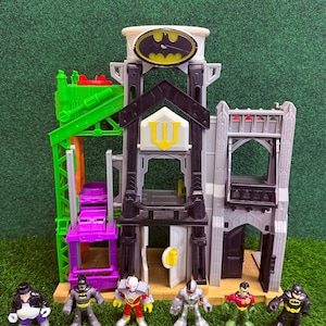 Peut inclure: Un ensemble de jeu Batman à plusieurs niveaux avec un Bat-Signal et une collection de figurines Batman. L'ensemble de jeu comprend un ascenseur vert, une plate-forme violette et un bâtiment noir et gris avec un emblème jaune. Les figurines incluent Batman, Robin et d'autres personnages.