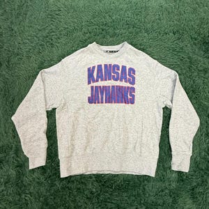 May include: A gray crew neck sweatshirt with the text "KANSAS JAYHAWKS" in blue and red block letters. The sweatshirt is made of a soft material and has long sleeves.