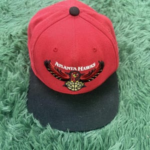 May include: A red baseball cap with a black brim and a black button on top. The front features an embroidered Atlanta Hawks logo with a hawk holding a basketball and the text Atlanta Hawks in white lettering across the centre of the design.