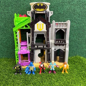 Peut inclure: Un ensemble de jeu sur le thème de Batman avec un bâtiment à plusieurs niveaux avec un Bat-Signal et un logo jaune et noir. Comprend Batman, Robin, Harley Quinn et d'autres figurines. L'ensemble de jeu a des éléments verts, violets, gris et noirs.