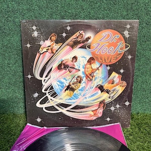 Puede incluir: Un álbum de vinilo vintage de Dr. Hook Live. La portada del álbum presenta una ilustración con temática espacial con miembros de la banda tocando la guitarra. El disco es negro y se ve la etiqueta de Capitol Records.