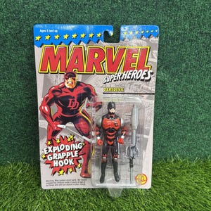 Może przedstawiać: Figurka Daredevil z Marvel Super Heroes w oryginalnym opakowaniu. Figurka jest czerwono-czarna, trzyma srebrną broń. Opakowanie zawiera logo Marvel i napis "Exploding Grapple Hook".