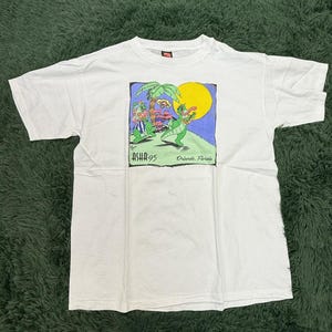 Peut inclure: T-shirt blanc avec un motif coloré d'un alligator de dessin animé jouant de la guitare, avec des palmiers et un soleil jaune. Le texte "ASHA 95 Orlando, Florida" est imprimé en dessous.