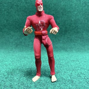 May include: A red action figure of a superhero, wearing a full body suit with a matching cowl and belt. The figure has a white emblem on the chest and is posed with fists clenched. The figure is standing on a green surface.