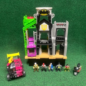 Peut inclure: Un ensemble de jeu sur le thème de Batman comprenant un bâtiment à plusieurs niveaux avec un Bat-Signal, un véhicule rose et noir, une grue verte et plusieurs figurines. L'ensemble de jeu est sur une surface verte.