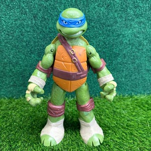 May include: A green action figure of Leonardo from Teenage Mutant Ninja Turtles. The figure has a blue mask, brown straps, and orange chest armor. It stands on a patch of green grass.
