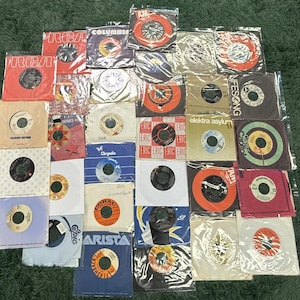 May include: A selection of vintage 45 RPM vinyl records in various sleeves. The sleeves display a range of colours, including red, orange, white, and blue, with text such as "RCA", "Columbia", and "Eric". The records are in plastic sleeves.
