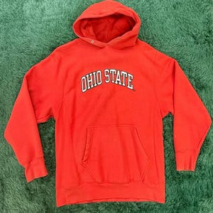 Puede incluir: Una sudadera con capucha roja con el texto "OHIO STATE" en letras blancas. La sudadera tiene un bolsillo delantero y una capucha con cordón. La prenda está hecha de un material suave y es adecuada para ropa casual.