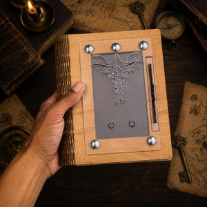 Puede incluir: Un diario rectangular de madera con lomo flexible de listones y acabado en color marrón claro. La cubierta frontal presenta un panel central de acrílico transparente asegurado por cuatro remaches metálicos plateados, con un diseño de cabeza de dragón grabado.
