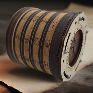 May include: A cylindrical wooden puzzle box featuring rotating rings with laser-engraved symbols including a skull, a rose, and a scorpion. The light-coloured wood is accented by dark brown rings and a circular faceplate secured with small metal screws.