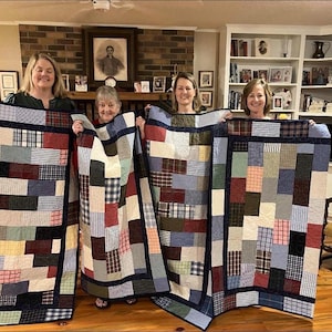 May include: Four handmade patchwork quilts, each with a unique arrangement of rectangular fabric patches in various colors and patterns, including plaid, stripes, and solid hues. The quilts are held up by four people.