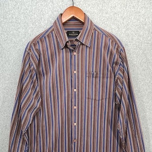 May include: A long-sleeved button-down shirt with vertical stripes in brown, blue, and burgundy. The shirt has a chest pocket and is hanging on a wooden hanger. The cuffs are rolled up.