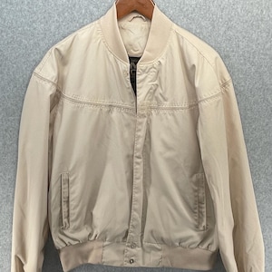 May include: A tan bomber jacket with a ribbed collar, cuffs, and hem. The jacket has a zipper closure and two front pockets. The jacket is hanging on a wooden hanger against a gray background.