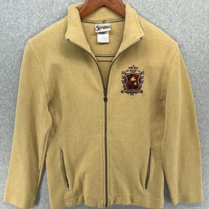 Peut inclure: Veste zippée moutarde avec un col montant et un petit écusson brodé sur la poitrine. L'écusson représente une couronne et un personnage de dessin animé. La veste a deux poches avant et est suspendue à un cintre en bois.
