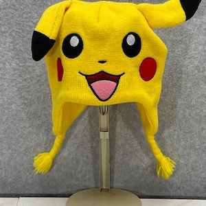 May include: A yellow knit hat shaped like Pikachu, featuring black-tipped ears, black eyes, red cheeks, and a smiling mouth with a pink tongue. The hat has yellow tassels and is displayed on a stand.
