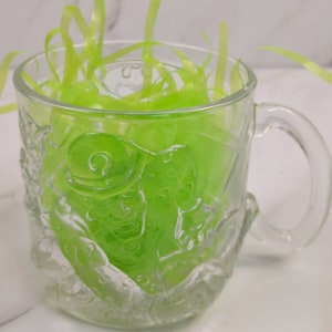 May include: A clear glass mug with a handle, filled with bright green shredded paper. The mug has an embossed design of a figure on the side. The mug is sitting on a white surface.