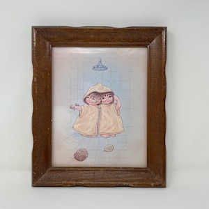 May include: A framed illustration of two children with a single head, wearing yellow hooded bathrobes, standing in a shower with a shower head above them. The illustration is set against a blue and white tiled background.