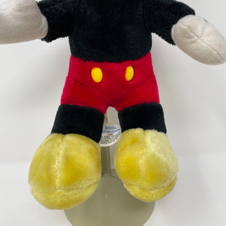 May include: A plush Mickey Mouse toy with black fur, red shorts with yellow buttons, and yellow shoes. The toy has a tag that reads "Disneyland Resort."