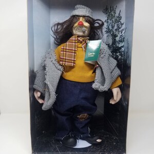 May include: A porcelain doll with a red nose and a gray hat, wearing a yellow sweater, a plaid scarf, and blue jeans with a patch on the knee. The doll is holding a pipe in its mouth and is standing in a box with a green background.