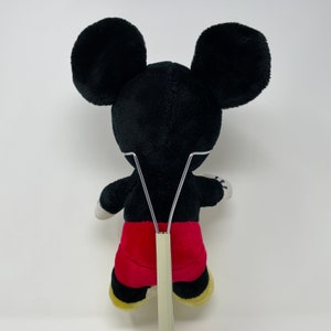 May include: A black and red plush Mickey Mouse toy with large ears. The toy is standing on a white stand.
