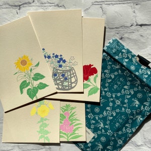 May include: Set of six blank note cards with floral illustrations. The cards are cream colored with a variety of flowers, including sunflowers, hibiscus, and bellflowers. The cards are packaged in a blue and white patterned bag.