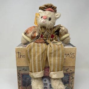 May include: A white plush cat wearing a brown and white striped overalls and a brown hat with a pink brim. The cat is sitting on a box with the words "The  ness" printed on it.