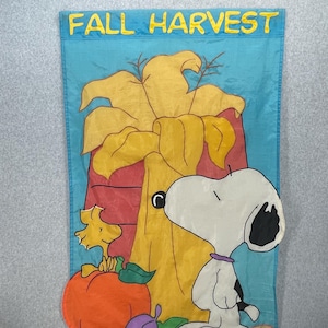 May include: A blue fall-themed flag with the words "FALL HARVEST" in yellow. The flag features Snoopy, Woodstock, a pumpkin, and a cornucopia. The Snoopy character is white with black ears and a black spot. The pumpkin is orange.