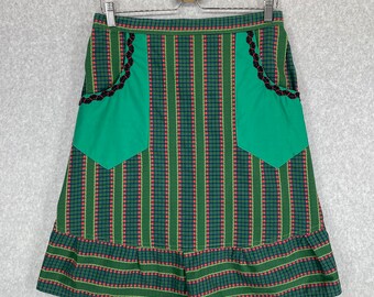 Pode incluir: Uma saia listrada verde e vermelha com dois bolsos verdes e acabamento preto. A saia tem uma bainha com babados.