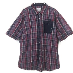 May include: A red, blue, and white plaid button-up shirt with a single chest pocket. The pocket has a black fabric overlay with the word "NATION" printed on it.