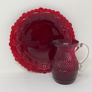 Può includere: Un'ampolla di vetro rossa con un motivo decorativo e un piatto di vetro rosso abbinato con un bordo smerlato.