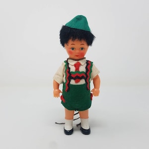 Puede incluir: Una pequeña muñeca de plástico que lleva un sombrero verde, una camisa blanca y un chaleco verde y negro con detalles rojos. La muñeca tiene el pelo negro y rasgos faciales pintados.
