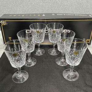 May include: Set of six clear crystal wine glasses with diamond-cut patterns. Each glass has a round base, a slender stem, and a flared bowl. The glasses are arranged on a dark surface, with a black box in the background.