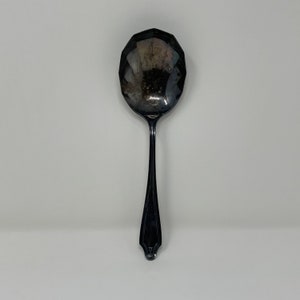May include: A silver serving spoon with a faceted bowl and a long, slender handle. The spoon has a decorative design at the end of the handle. The spoon is set against a white background.