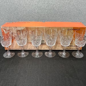 May include: Set of six clear crystal wine glasses with intricate cut patterns. The glasses are arranged in front of a vintage box with text. Each glass has a round base and a slender stem.