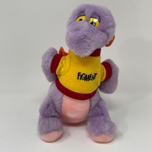 Peut inclure: Dragon en peluche violet Figment portant un t-shirt jaune avec des bordures rouges et le mot "Figment" imprimé sur le devant.