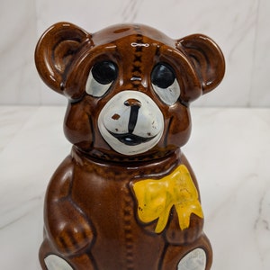 以下が含まれることがあります： 黄色のリボンが付いた茶色のセラミック製のテディベア貯金箱。クマは黒い目と白い鼻と口をしています。
