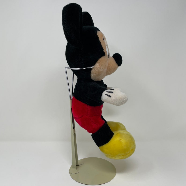 May include: A black and red plush Mickey Mouse toy sitting on a metal stand. The toy has yellow shoes and is in a sitting position.
