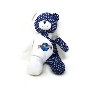 May include: A plush teddy bear with a split design. One side is white, the other is navy blue with white stars. The white side features the "Universal Studios" logo. The bear has a small white bow.