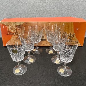 May include: Set of six clear crystal wine glasses with diamond-cut patterns. The glasses have a stemmed base and a wide bowl. They are arranged in front of an orange box with text and images of glasses.