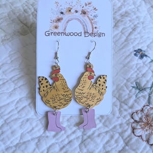May include: Wooden earrings with a chicken design. The chickens are tan with black spots and red combs, and are wearing pink wellington boots. The earrings hang from silver hooks. The card behind the earrings says "Greenwood Design".