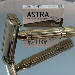 May include: A gold-toned metal double edge safety razor with a textured, knurled handle. Behind the razor sits a white and brown cardboard box labelled ASTRA SUPERIOR PLATINUM DOUBLE EDGE with a graphic of a razor blade and the number 5.